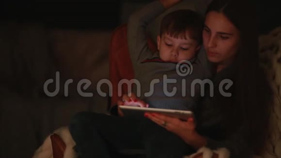 美丽的母亲坐在椅子上他的家和儿子搭在腿上在触摸屏上看动画片视频的预览图