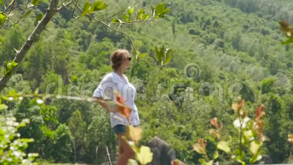 年轻女子站在背景上夏天的山覆盖着绿色的森林女孩在欣赏青山美景视频的预览图