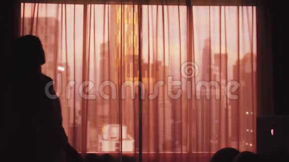 年轻女子的剪影拉开了大窗户上的窗帘张开双臂望着城市上的公寓视频的预览图