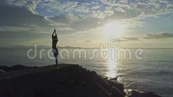 女孩剪影瑜伽在岩石上对抗海洋上空升起的太阳视频的预览图