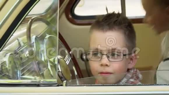 小男孩开着那辆站立的复古车在摄像机上嬉戏穿复古衣服的男孩坐在美丽的米色里视频的预览图