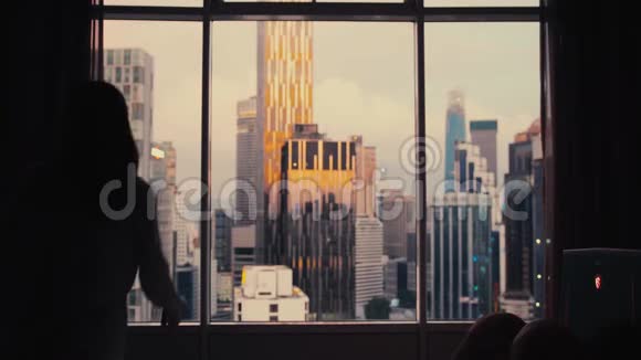 年轻女子在城市建筑背景下揭开公寓的大窗户城市的日落慢动作视频的预览图