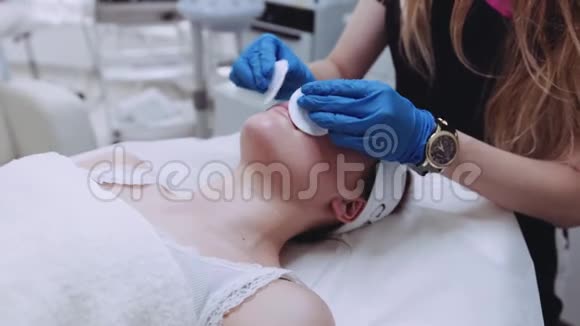 一个漂亮的女人闭着眼睛躺在美容师桌子上皮肤科医生用棉花海绵做手术视频的预览图