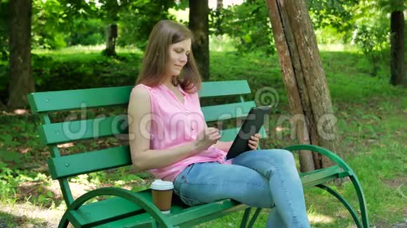 漂亮女孩在公园里坐在长凳上网上买东西快乐的女人在露天通过网络平板电脑购物视频的预览图