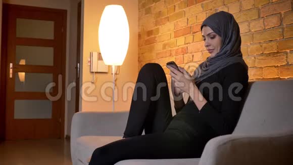 在室内沙发上坐着的穆斯林成年女性戴着头巾在手机上发短信视频的预览图