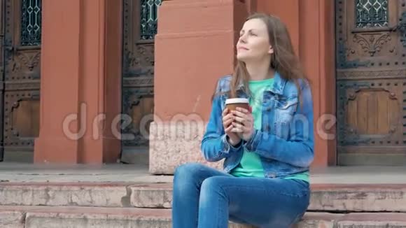 漂亮的女孩穿着棉袄坐在台阶上喝咖啡背景中的老式建筑视频的预览图