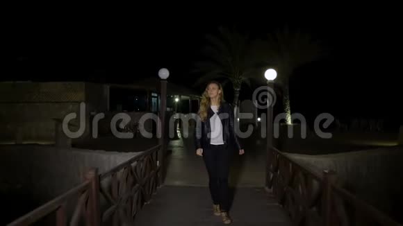 一位身穿皮夹克的金发碧眼年轻女子独自走在灯柱间的木人行桥上或在晚上或晚上视频的预览图