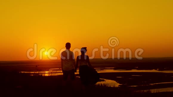 在夕阳下可爱的一对情侣年轻的男人和美丽的女孩站在夏日的草地上望着阳光剪影视频的预览图