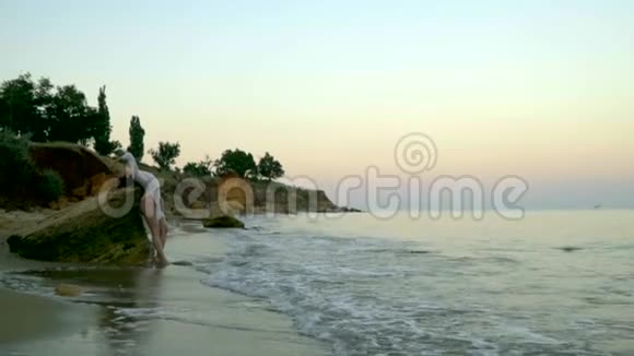 一个细长的金发碧眼穿着一件漂亮的白色连衣裙站在海滩边风景如画的海湾里的岩石上视频的预览图