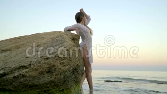 一个穿着白色短裙的金发女郎站在岩石旁边看着大海上的夕阳视频的预览图