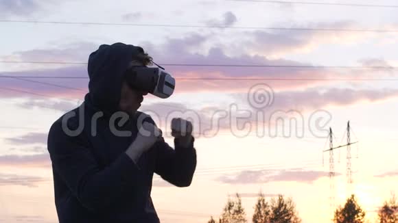 青年拳击手在VR360耳机训练中在日落时踢虚拟现实战斗中的剪影慢动作视频的预览图