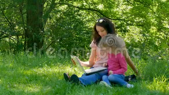 美丽的小女儿和她的年轻母亲坐在草地上的城市公园与白色笔记本电脑看着视频的预览图