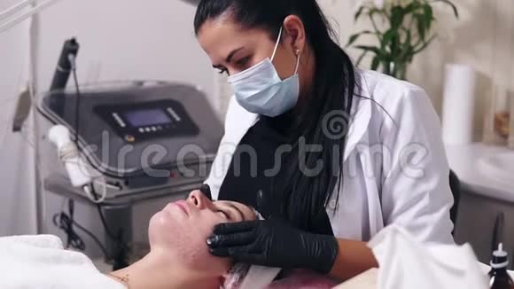 专业美容师正在为躺在上面的皮肤有瑕疵的年轻女性涂抹面霜并做面部按摩视频的预览图
