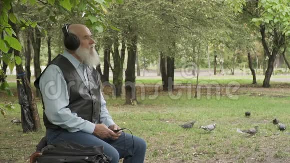 白发老人坐在公园的长凳上听音乐戴着耳机慢镜头视频的预览图