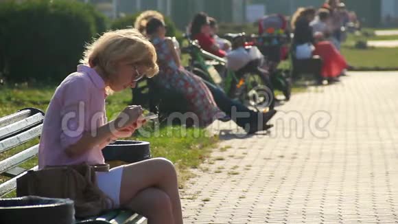 美丽的年轻女子在公园的长凳上用智能手机敲击触摸屏视频的预览图