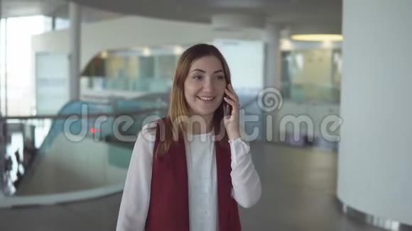 穿着优雅衣服的积极的年轻女孩在机场的智能手机上讲话视频的预览图