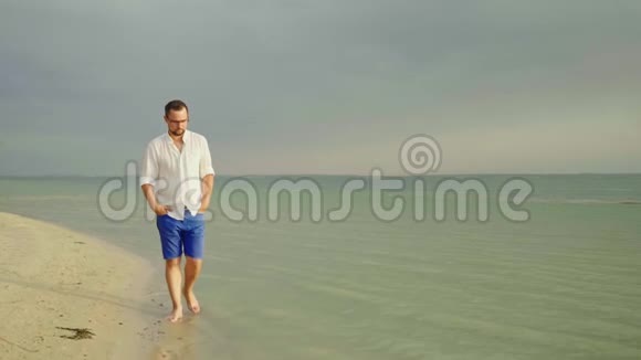 一个年轻漂亮的男人穿着浅色的衣服和眼镜沿着海边走摆脱忧虑自由视频的预览图