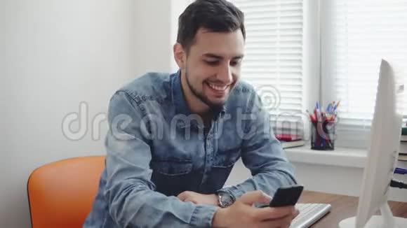 年轻快乐的上班族在办公室里用手机坐在桌旁端着电脑电话和杯子微笑着视频的预览图