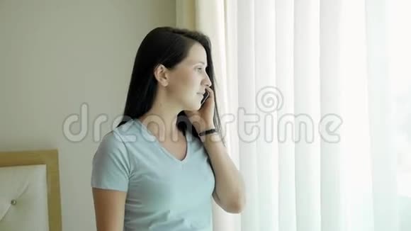 年轻女性使用智能手机打电话的肖像白人模特站在窗口拿着手机说话视频的预览图