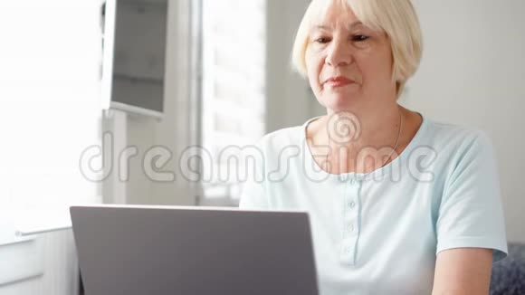 年长的金发女人在家里用笔记本电脑工作收到好消息兴奋和高兴视频的预览图