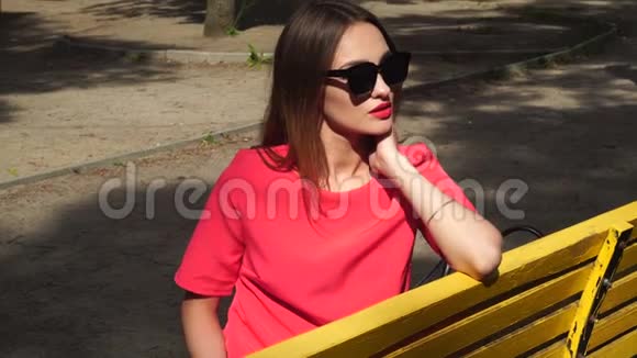 迷人的年轻女士穿着红色西装戴着太阳镜坐在公园的长凳上视频的预览图