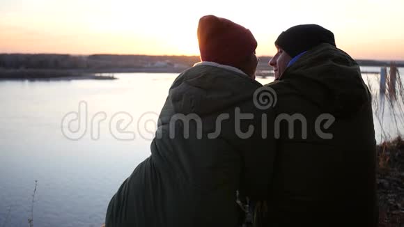一对可爱的夫妇穿着夹克和帽子坐在河岸上欣赏日落和亲吻慢动作1920x1080视频的预览图