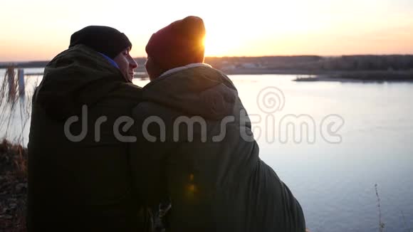 在河岸上一对相爱的夫妇穿着夹克和帽子坐在日落背景上有镜头效果慢动作视频的预览图