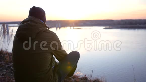 一个穿着夹克和帽子的年轻人坐在桥附近的河岸上欣赏美丽的日落慢动作视频的预览图