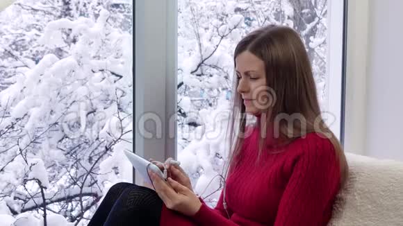 漂亮的女孩坐在窗台上看着窗户用带耳机的平板电脑冬天在外面视频的预览图