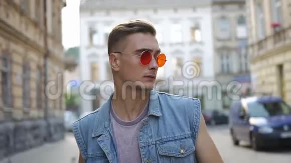 戴着红墨镜的男人脱下牛仔裤背心穿上他的肩膀沿着街走视频的预览图