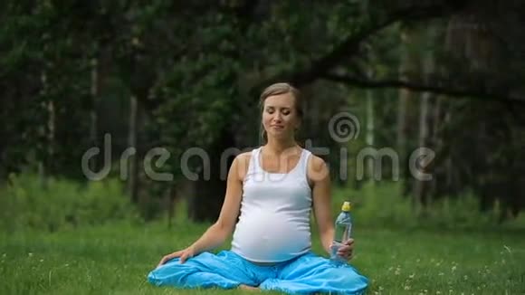 孕瑜伽女饮水从一瓶在莲花位置公园草地户外森林视频的预览图