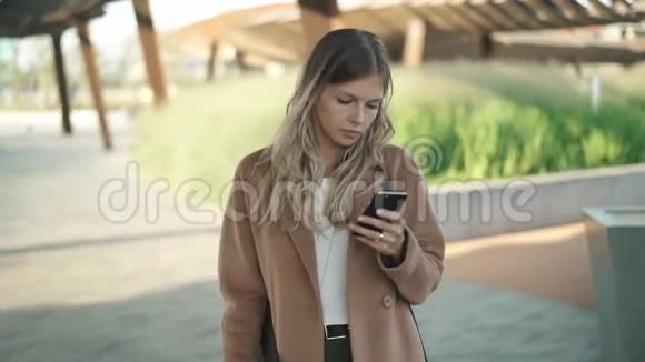 秋天穿着大衣的金发美女在散步看着自己的智能手机视频的预览图