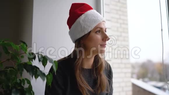 美丽的年轻女子戴着圣诞老人的帽子坐在窗台上端着茶和看书期待着视频的预览图