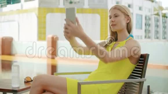 一个穿着黄色衣服的女人坐在游泳池附近的椅子上用她的智能手机自拍视频的预览图
