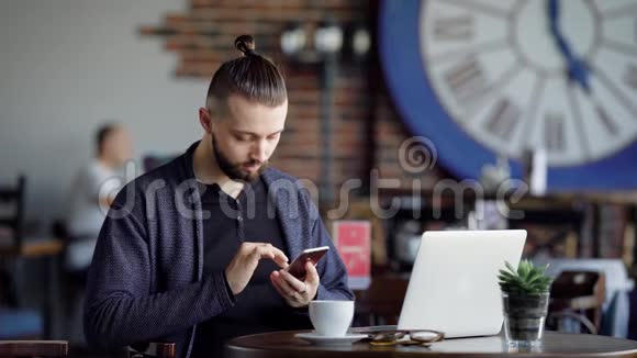一个成年人在一家现代化的咖啡馆里喝咖啡一个头发扎在尾巴上的男人浏览着视频的预览图