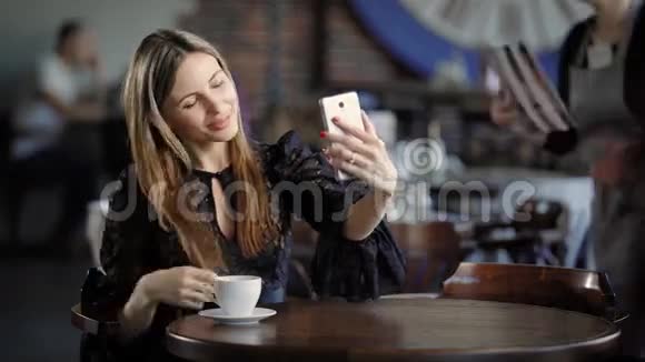 年轻女子坐在咖啡馆里用现代智能手机自拍穿着黑衣的女士看着视频的预览图