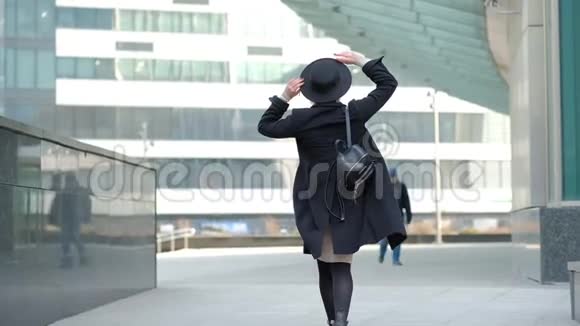 女人玩现代黑色帽子移动宽边起首在黑色外套底部在街道慢动作低角度拍摄视频的预览图