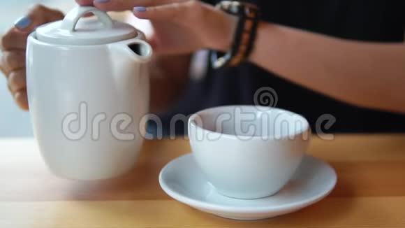 在咖啡馆的木桌上用白瓷杯从茶壶里倒热茶的女性手的近景视频的预览图