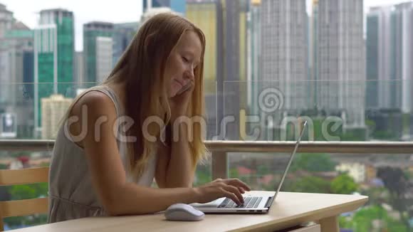 年轻女性自由职业者在阳台上的笔记本电脑上工作背景是一个充满摩天大楼的市中心她是视频的预览图