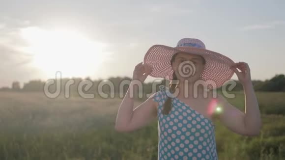 一个穿着浅色连衣裙和帽子的快乐女人的肖像日落时分女孩带着谷类植物走在田野上视频的预览图