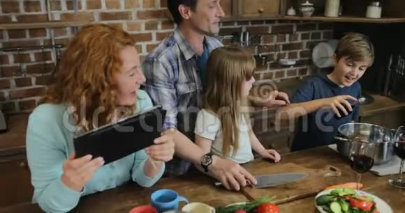 一家人在厨房里用平板电脑的食谱一起做饭父母带着两个孩子在视频的预览图