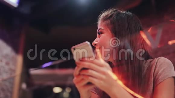 年轻迷人的女人美丽的微笑是使用智能手机在晚上坐在酒吧或餐馆里的女孩视频的预览图