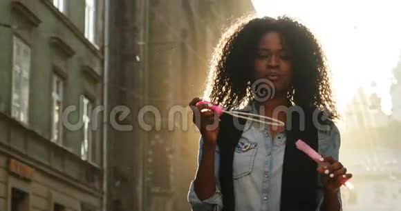 精力充沛的美国黑人女性卷发在阳光明媚的日子里吹着街道上的肥皂泡视频的预览图