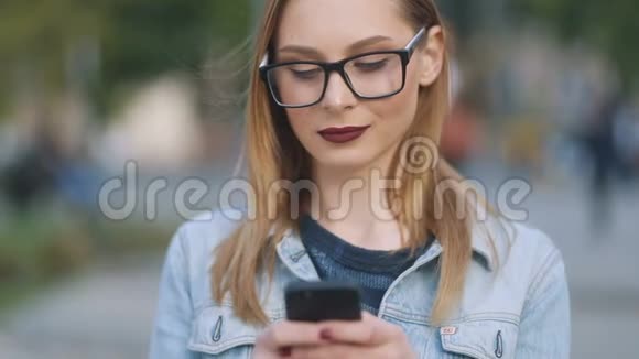 一位戴着眼镜的美女在城市的智能手机上发短信时她正为照相机微笑视频的预览图