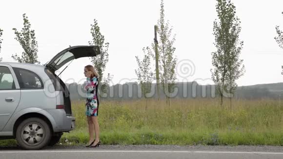 一个金发女孩在一条乡间小路上开着后备箱的破车旁惊慌失措地打着手机视频的预览图