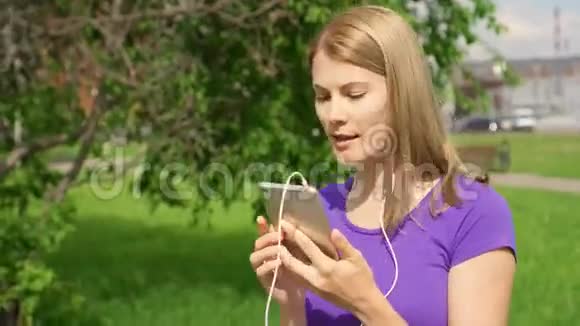 女人在公园里用耳机听手机音乐用她的智能手机跳舞唱歌视频的预览图