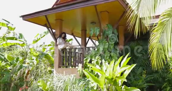 在热带森林的别墅里年轻女子坐在阳台上使用手机漂亮女孩在户外上网聊天视频的预览图