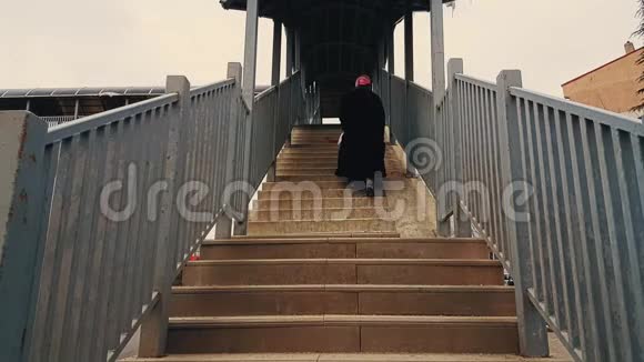 穿着黑色外套和头巾的老妇人走上人行天桥的楼梯视频的预览图