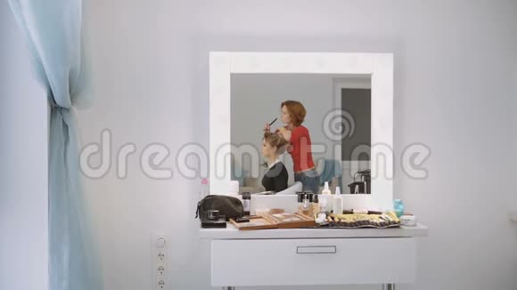 豪华美容院的女理发师正在与年轻的金发模特站在一起工作从镜子里看到视频的预览图