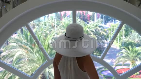 一个戴着白色大帽子和白色连衣裙的女人站在一扇圆圆的大窗户附近欣赏着它的本质4ka慢动作视频的预览图